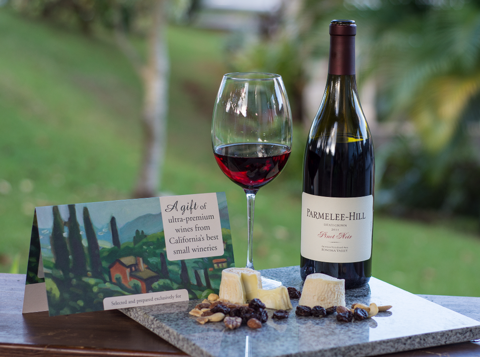 a glass of red wine next to a bottle upon a cheese and nut board by a gift card